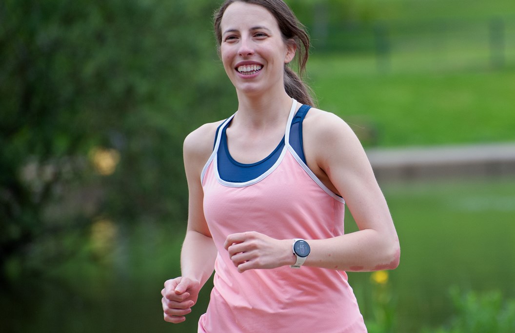 Emily, a woman in her twenties with brown hair, running towards the camera while smiling. She is wearing a salmon pink running vest and black shorts. Immediately behind her is a lake, with a grassy embankment and green trees in the background.