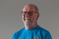 A man with a white beard and glasses, wearing a blue t-shirt with a motif on the left breast, stands smiling at the camera with his right hand placed over his left hand in front of his torso.