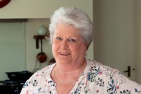 A woman (Anna) in a white, flowery top, stood in a kitchen, smiling at the camera, holding a plate of cooked chicken strips, salad and sauce with her right hand. Her left hand is on her hip.