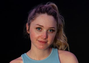 A young woman wearing a pale blue top and shorts, facing the camera with her arms folded and smiling slightly.