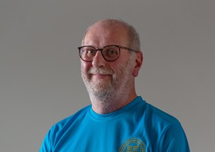 A man with a white beard and glasses, wearing a blue t-shirt with a motif on the left breast, stands smiling at the camera with his right hand placed over his left hand in front of his torso.