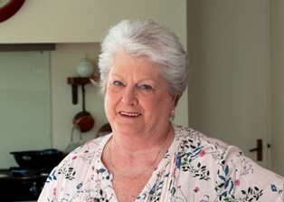 A woman (Anna) in a white, flowery top, stood in a kitchen, smiling at the camera, holding a plate of cooked chicken strips, salad and sauce with her right hand. Her left hand is on her hip.