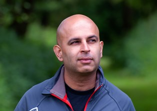 A man in the foreground looking up towards the sky as he walks in woodland with green trees and bushes behind him.