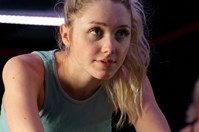 A young woman wearing a pale blue top and shorts pedalling on an exercise bike in a gym, with two more women pedalling in the background.