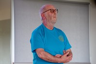 A man with a white beard and glasses, wearing a blue t-shirt with a motif on the left breast, stands facing to the right with his arms in front of him as if cradling something.