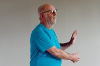 A man with a white beard and glasses, wearing a blue t-shirt, faces to the right, with his left palm raised and his right arm stretched out in front of him and to the left.