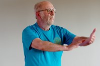 A man with a white beard and glasses, wearing a blue t-shirt with a motif on the left breast, stands facing the right with his left arm stretched out in front of him, with the palm facing inwards, and his right arm outstretched with the palm flat.