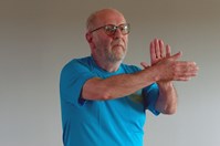 A man with a white beard and glasses, wearing a blue t-shirt with a motif on the left breast, stands facing forwards with his arms outstretched in front of him and his palms touching. His left palm is facing outwards and his right palm is facing inwards.