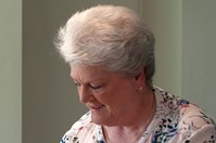 A woman (Anna) in a white, flowery top, standing behind a worktop in a kitchen. She is using tongs to serve cooked chicken strips up onto a plate.