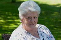 A lady (Anna) in a white, flowery top and black trousers, sat in a garden chair next to a table, while smiling at the camera. She is sat on a lawn with bushes behind her. It is a sunny day.