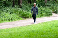 A man, some distance away, wearing jeans and a grey zip up jacket, walking along a gravel path in front of several tall trees