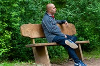 A man in a grey zip up jacket, jeans and trainers sitting on a wooden bench, looking right. He is sat in front of green bushes and trees.