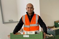 A man in an orange high visibility jacket and jeans holding a green cardboard box full of food donations. He is stood in front of a stack of similar boxes in a small hall.