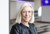 A lady with light hair and glasses is stood smiling at the camera. She is wearing a black leather jacket over a blue top, with a silver necklace. She is stood in front of a blurry backdrop of a conference hall.